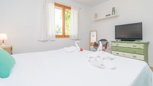 A hotel room with a bed, a television, a dresser, a window with curtains, and a small shelf with items.