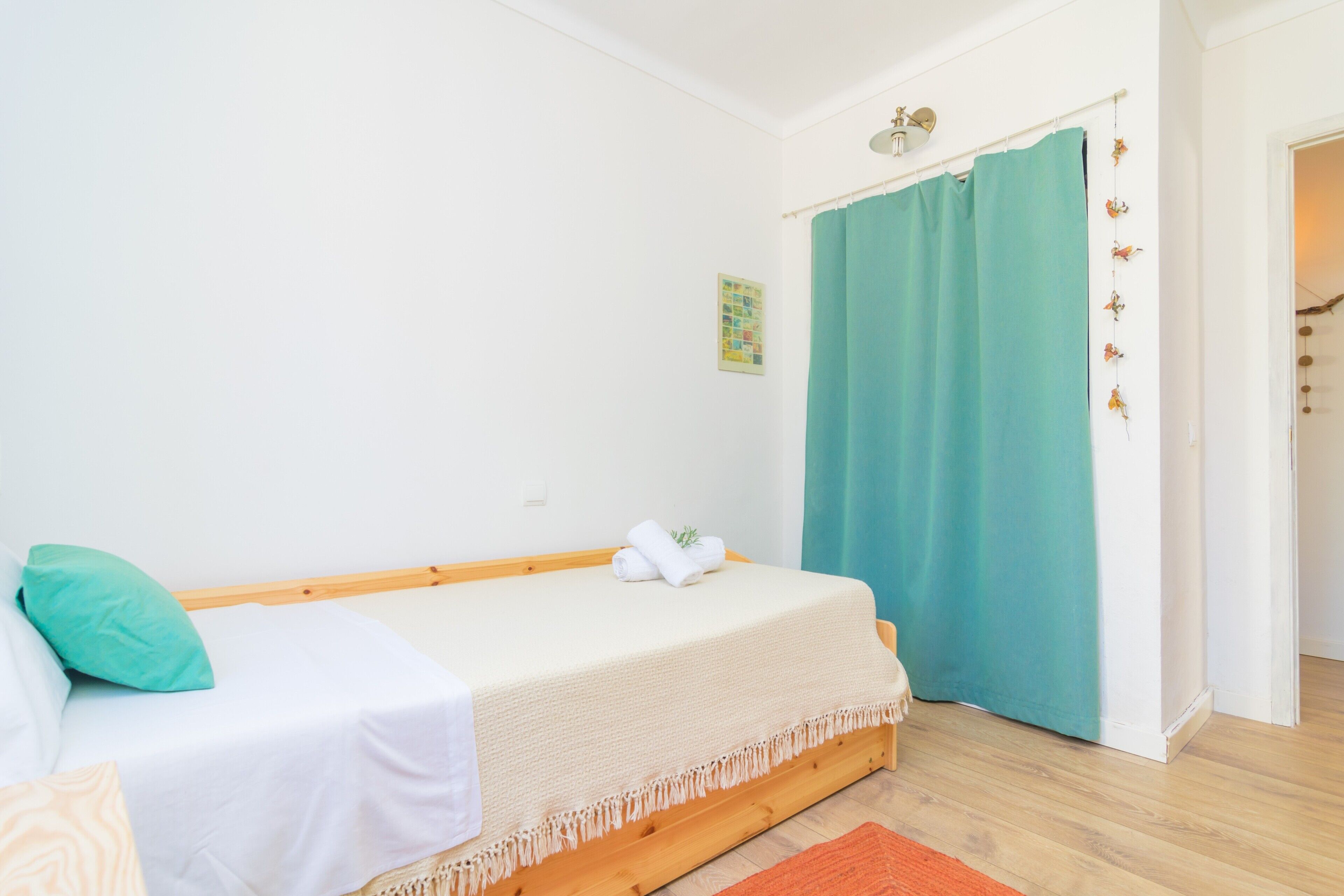 Een enkel bed met een wit matras en een beige deken, een blauwgroen gordijn en een houten vloer.