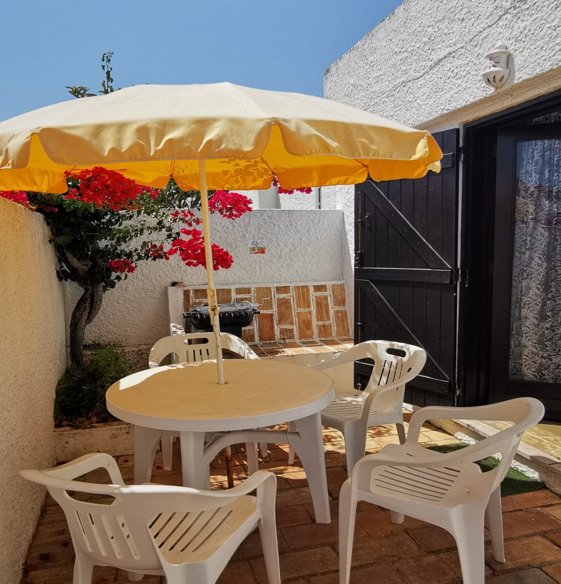 An outdoor patio with a round table and chairs, a yellow umbrella, and red flowers.