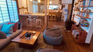 A cozy living room with a sofa, ottoman, and coffee table, a kitchen area with wooden cabinets, and a bookshelf filled with books and toys. - Guesthouse SORA - Hostel (Minamiizu)