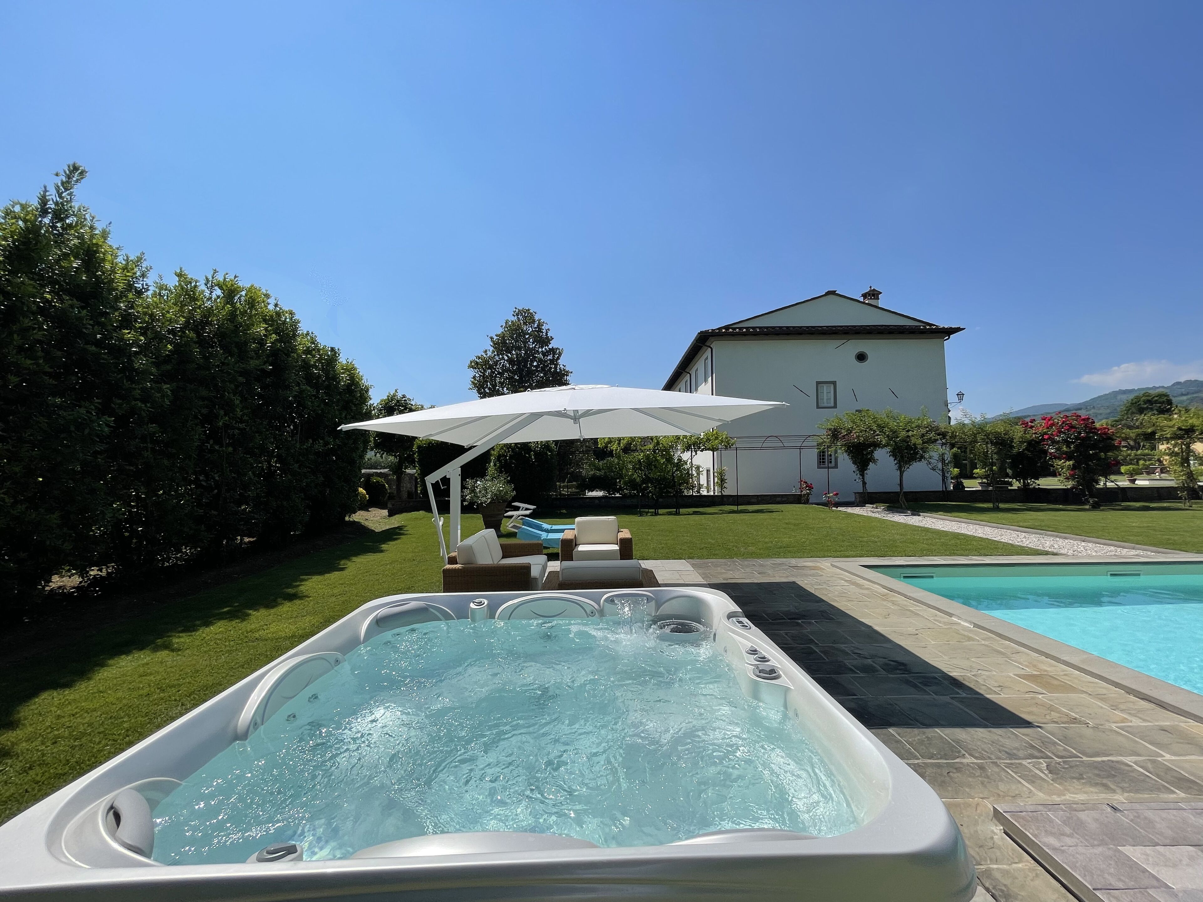 A hot tub with a white umbrella, a pool, and a house in the background.