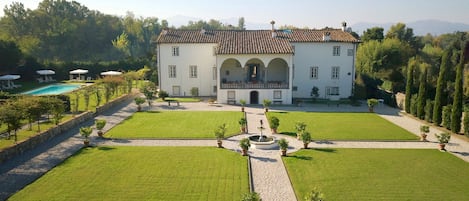 Un edifici gran i de color blanc amb teulada enrajolada, envoltat de jardins ben cuidats i una piscina.