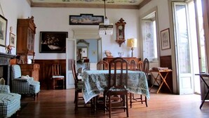 Une salle à manger avec une table en bois, des chaises et un lustre. On y trouve une cheminée avec un manteau de cheminée et une horloge au mur.