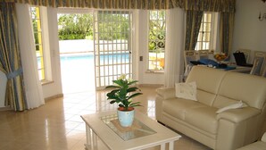 A living room with a beige sofa, a glass coffee table with a potted plant, and a view of a swimming pool through large windows. - Beautiful 4 bedroom villa in the Alto Golf (ALVOR)