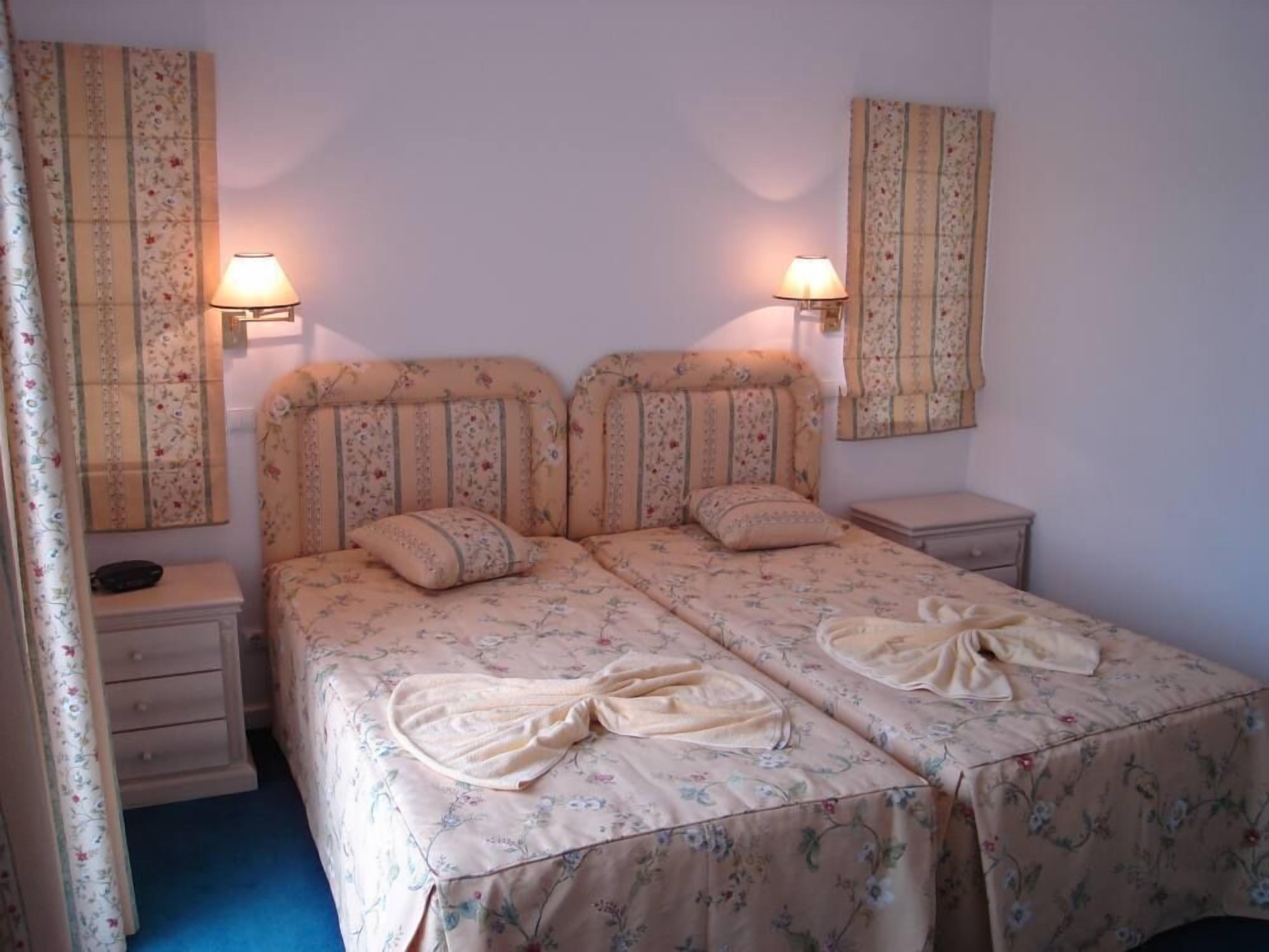 Un llit de matrimoni amb roba de llit floral, dues taules de nit i dues làmpades penjades a la paret.
