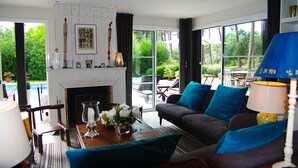 Ein modernes Wohnzimmer mit Kamin, einer blauen Kissen-Sofaecke, einem Essbereich mit Tisch und Stühlen und großen Fenstern mit Blick ins Freie.