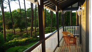 Eine Holzterrasse mit einem Korbstuhl, umgeben von einem weißen Geländer und Blick auf eine bewaldete Gegend.