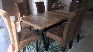 A dining area with a wooden table and cushioned chairs, a kitchen in the background, and a painting on the wall.