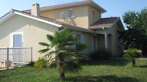 A two-story house with a tiled roof, a satellite dish, and a small garden with palm trees.