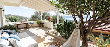 A covered outdoor seating area with a view of the sea, a hammock, and a barbecue grill.