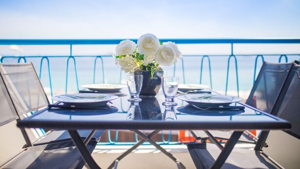 Mesa para dos con un jarrón de flores blancas, copas y platos, con vista a una barandilla azul y al océano.