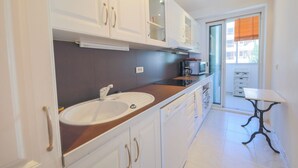 Una cocina moderna con armarios blancos, un lavamanos y una encimera. Hay una ventana con persianas y una pequeña mesa con una silla.