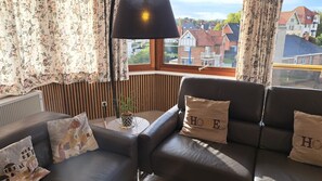 A room with a black leather sofa, a small table with a plant, and a window with a view of houses. - Apartment/ flat - (Coxyde)
