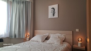 Una cama bien hecha con ropa blanca, un cabecero y dos lámparas de noche. Un cuadro enmarca una obra de arte en la pared sobre la cama.