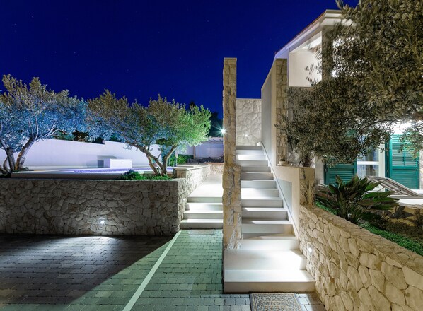 A nighttime view of a modern building with a staircase leading to a second floor, surrounded by stone walls and illuminated trees.