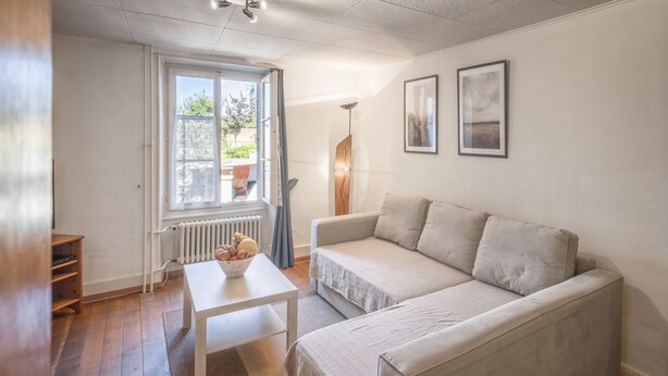 A small, well-lit room with a white sofa, a small table with a bowl of fruit, and a radiator. There is a window with a view of greenery outside.