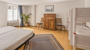 A bedroom with a bed, a wooden wardrobe, two chairs, a potted plant, and a window with curtains.