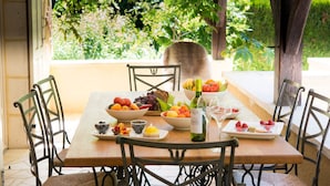 Une table à manger dressée avec des fruits, une bouteille de vin et des pâtisseries, entourée de chaises, et offrant une vue sur la verdure à l’extérieur.