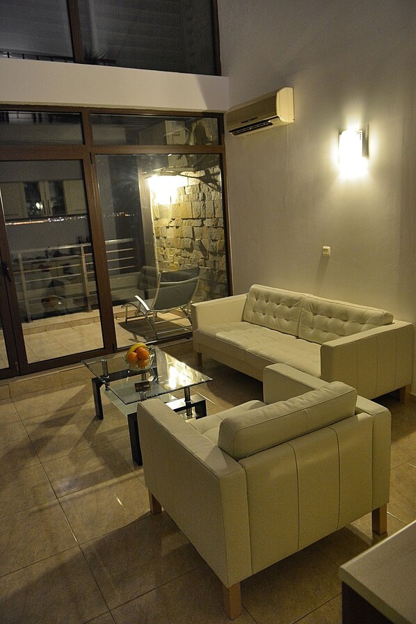 Una sala de estar moderna con un sofá, un sillón y una mesa de centro de vidrio. Se aprecia una pared de piedra y un equipo de aire acondicionado en la pared.