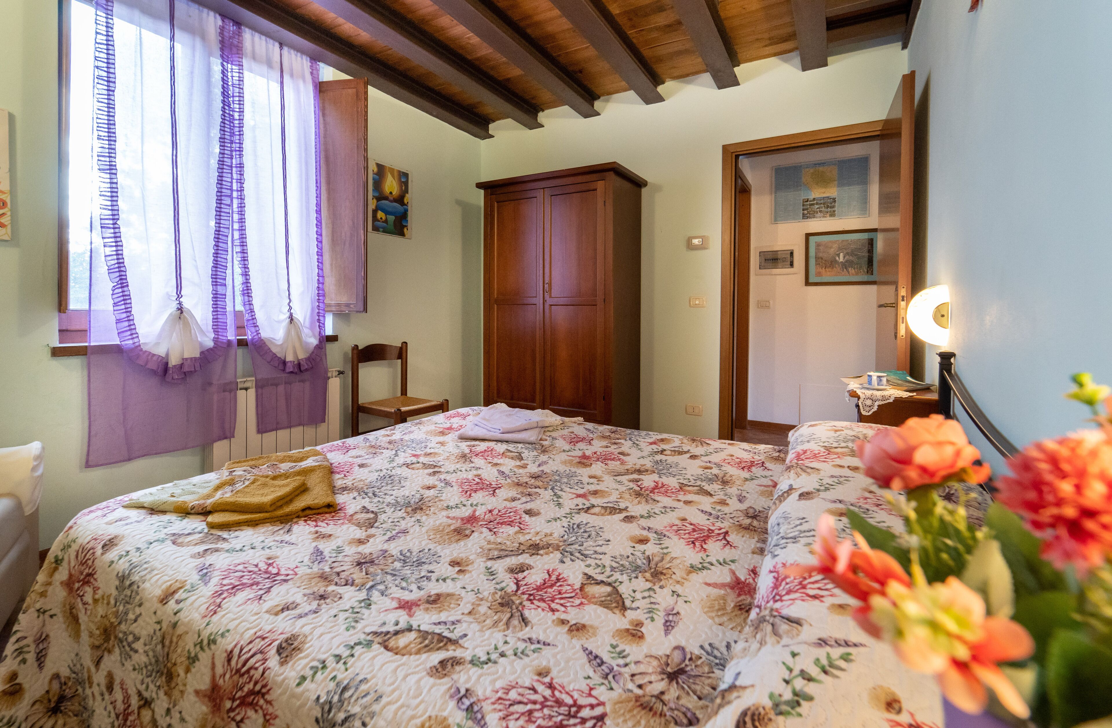 Un dormitorio con colcha estampada, un armario de madera, una ventana con cortinas moradas y una mesita de noche con lámpara.