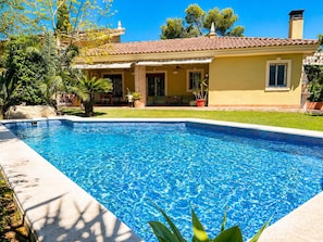 Une piscine dont le bord est recouvert de carreaux, entourée d’une pelouse et bordée d’un bâtiment dont le toit est également recouvert de carreaux.