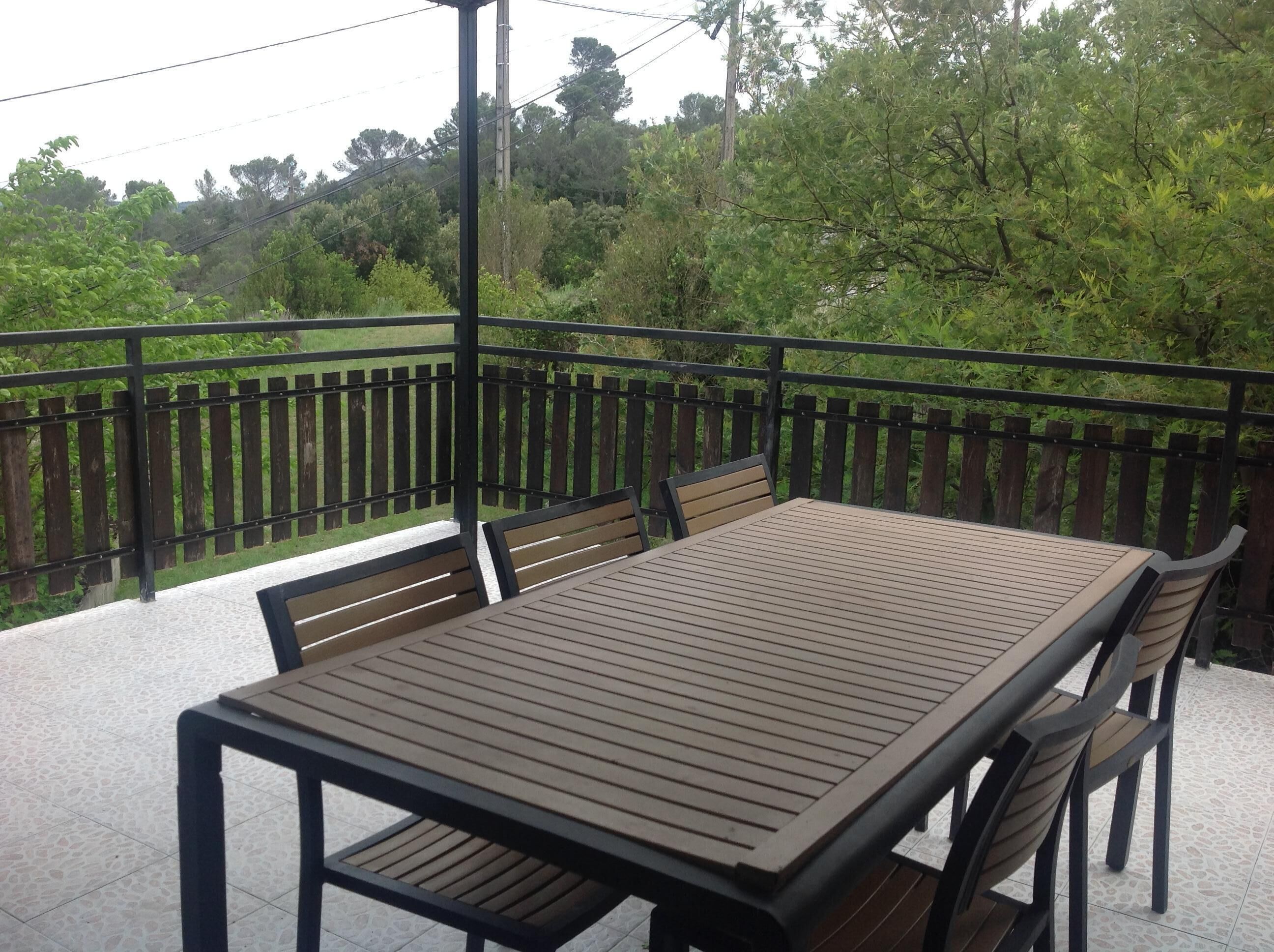 Uma mesa e cadeiras de madeira em uma varanda com guarda-corpo de metal, com vista para uma paisagem verdejante.