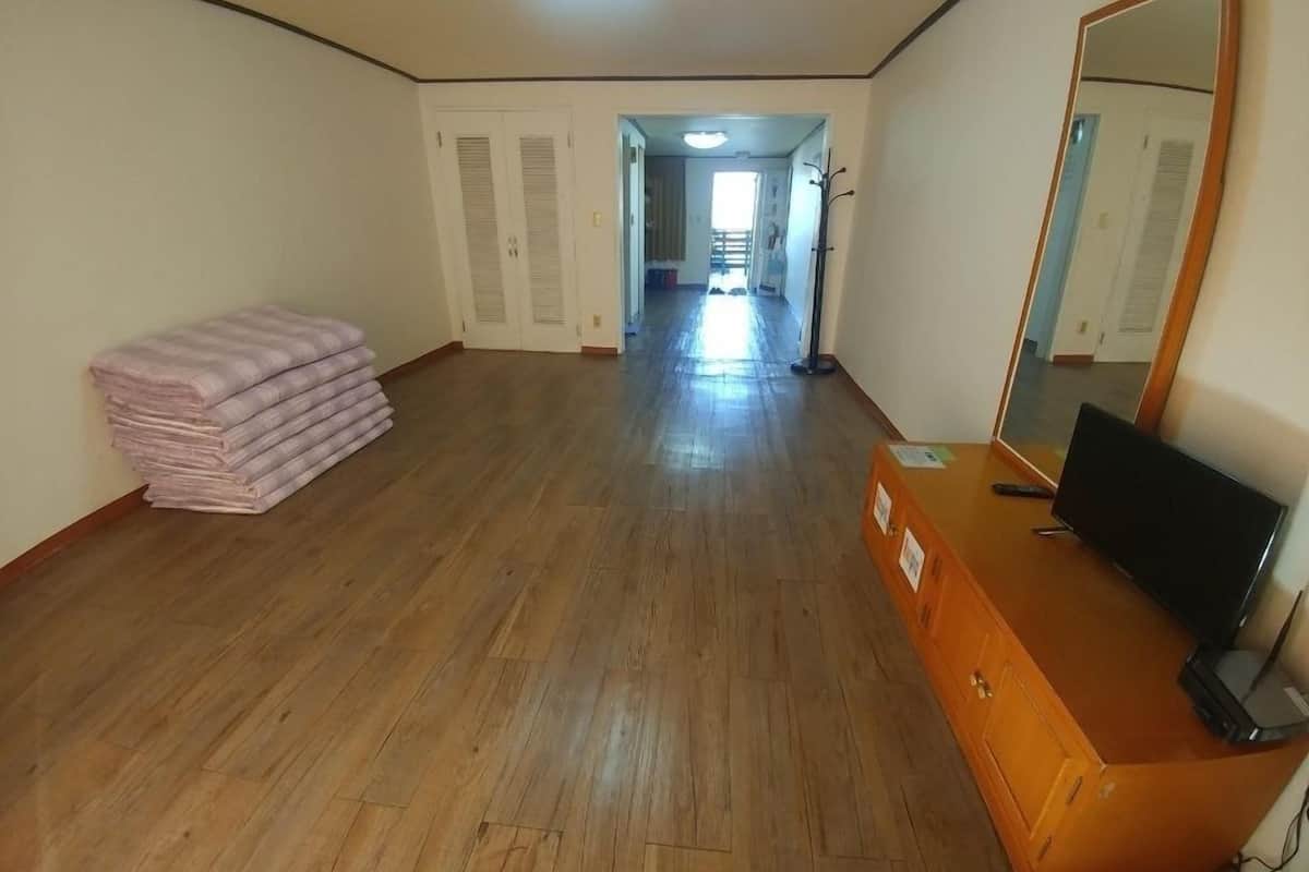Una habitación con piso de madera, un montón de mantas dobladas, un aparador con espejo y un televisor.
