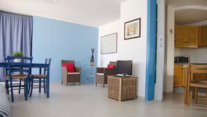 A living room with a blue wall, a dining area with blue chairs, a TV on a wooden stand, and a kitchen visible through an open door. - Casa Palme, APARTMENT AT SEA (Marzamemi)