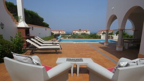 Une terrasse dotée de chaises longues blanches et d’une petite table, offrant une vue sur une piscine et sur la mer.