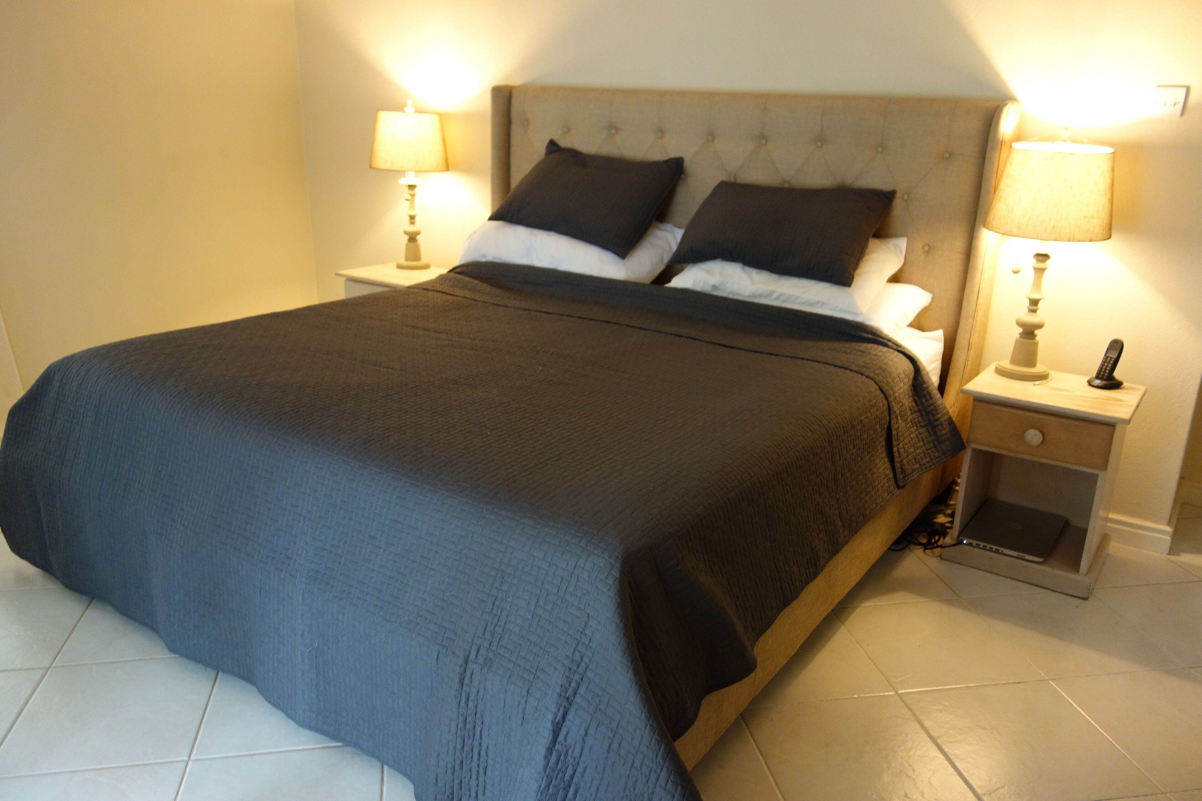 A neatly made bed with a dark blue bedspread, two bedside lamps, and a nightstand with a remote control.