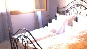 Una cama con estructura metálica y ropa de cama blanca en una habitación con ventana, espejo y un cuadro en la pared.