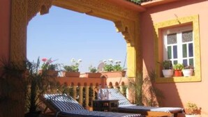 Un espacio al aire libre iluminado por el sol con dos sillones de descanso y una pequeña mesa, rodeado de macetas con plantas y un arco decorativo.