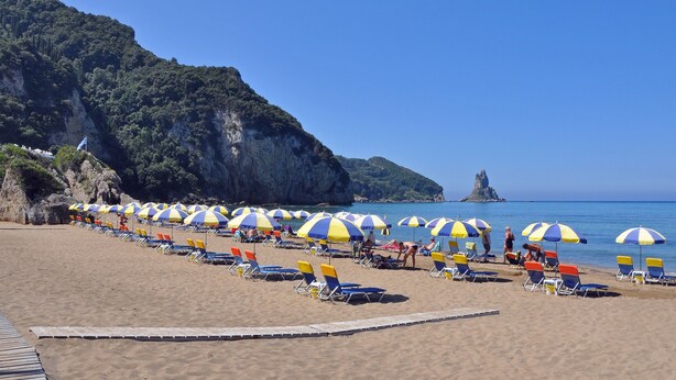 Ein Strand mit Reihen von Liegestühlen und Sonnenschirmen, einer Holzpromenade und einer felsigen Klippe im Hintergrund.