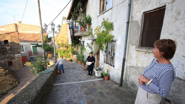 Une ruelle étroite pavée de pierres, avec des bâtiments de chaque côté, un enfant qui joue au ballon et une femme qui se tient sur le trottoir.