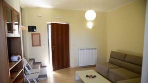 Un salon avec un canapé, une table basse et un téléviseur. Une porte donne sur une autre pièce et un radiateur est fixé au mur.