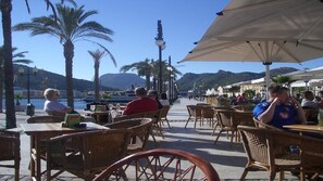 Ein Straßencafé mit Korbstühlen und -stühlen, Blick auf das Meer und Berge im Hintergrund.
