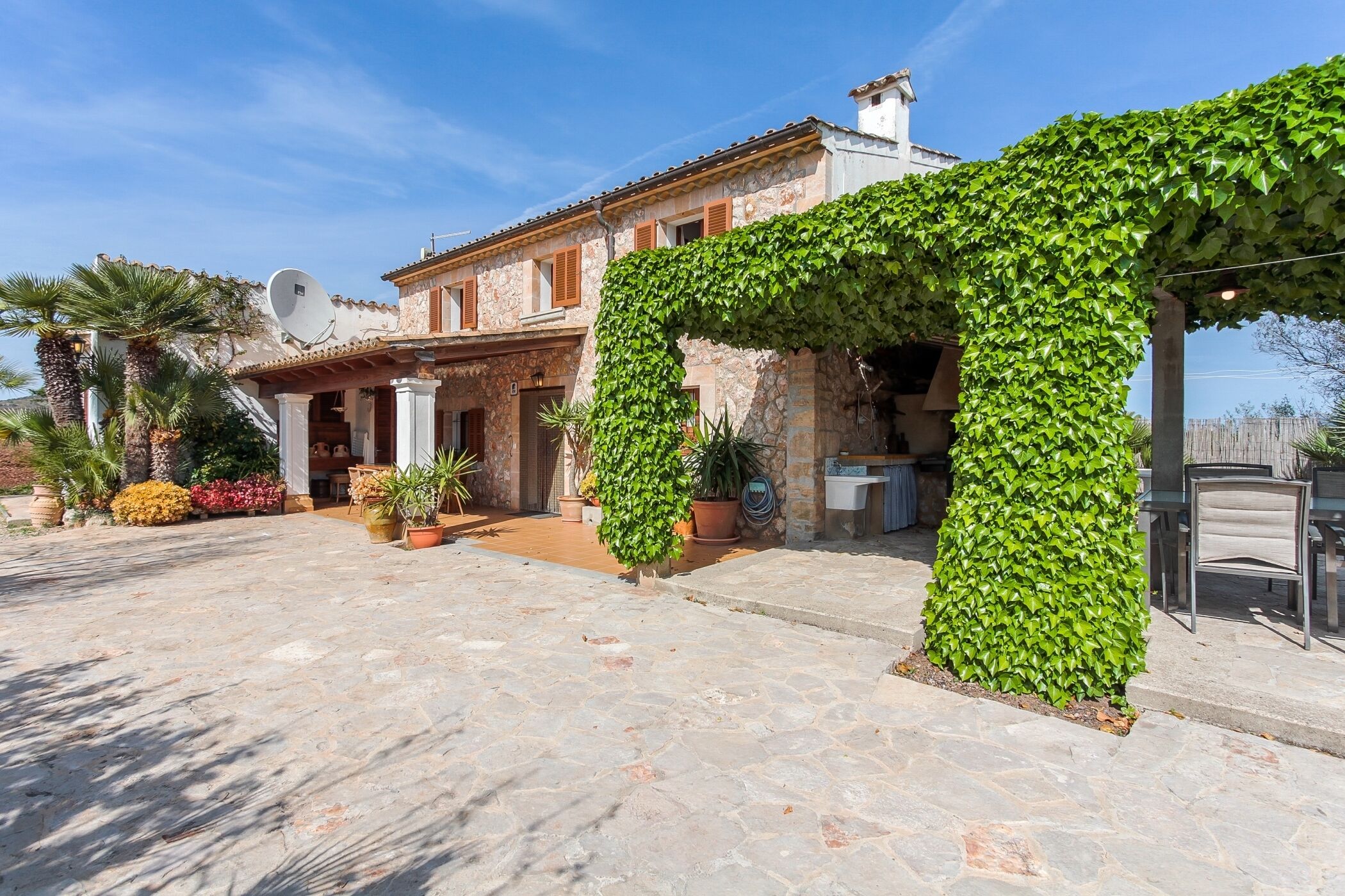 A stone building with a satellite dish, potted plants, and a pergola covered in ivy.