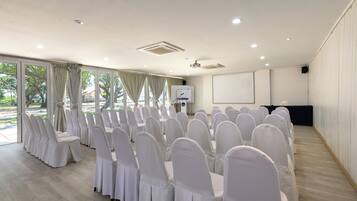 A conference room with rows of white-covered chairs and tables, large windows with curtains, a whiteboard, and a projection screen.