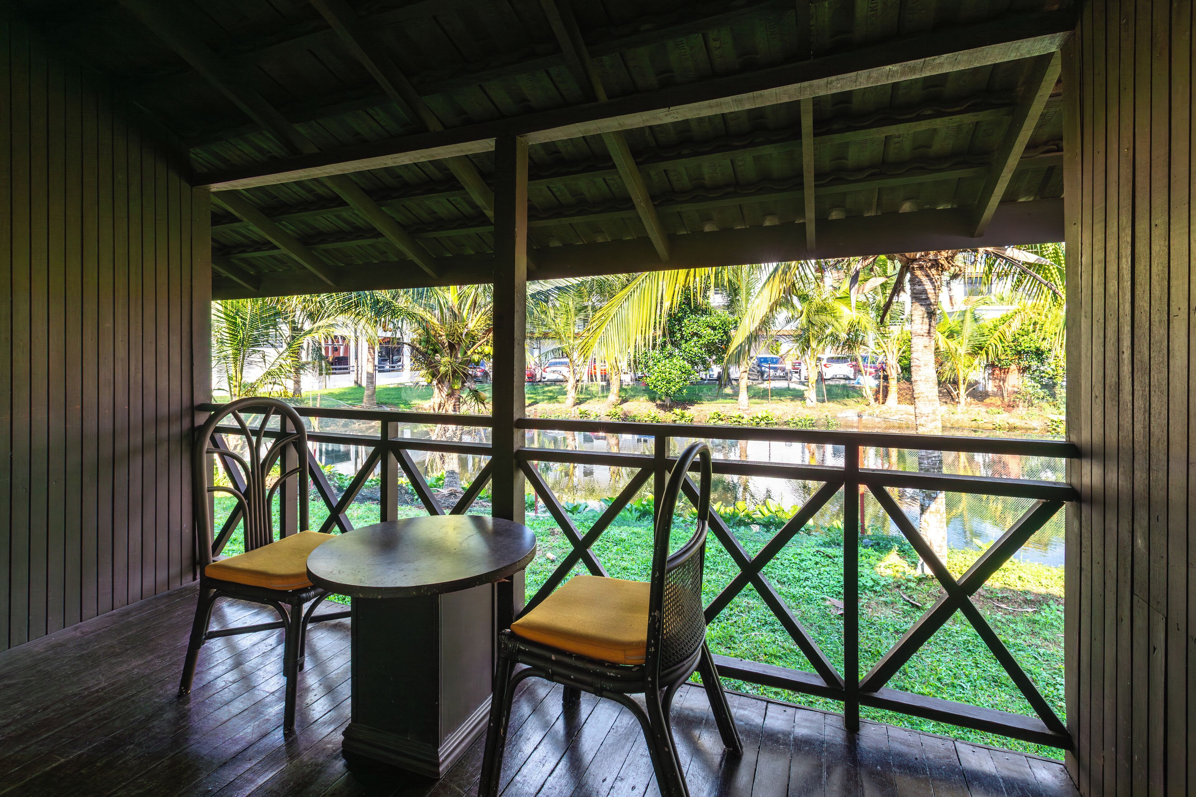 A covered outdoor seating area with a table and two chairs, overlooking a garden with palm trees and parked cars.