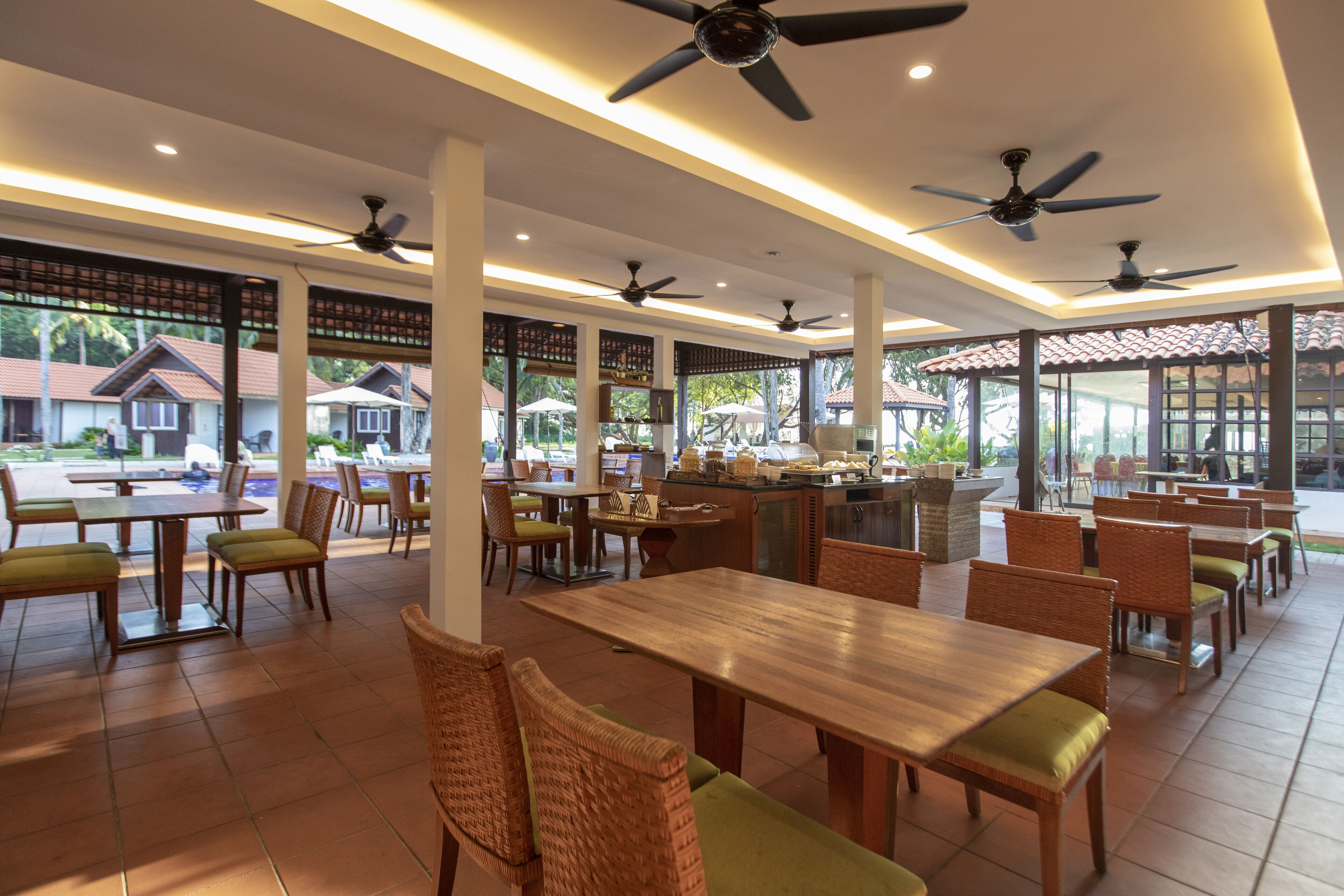 Um restaurante espaçoso com várias mesas e cadeiras, ventiladores no teto e grandes janelas que oferecem vista para o exterior.