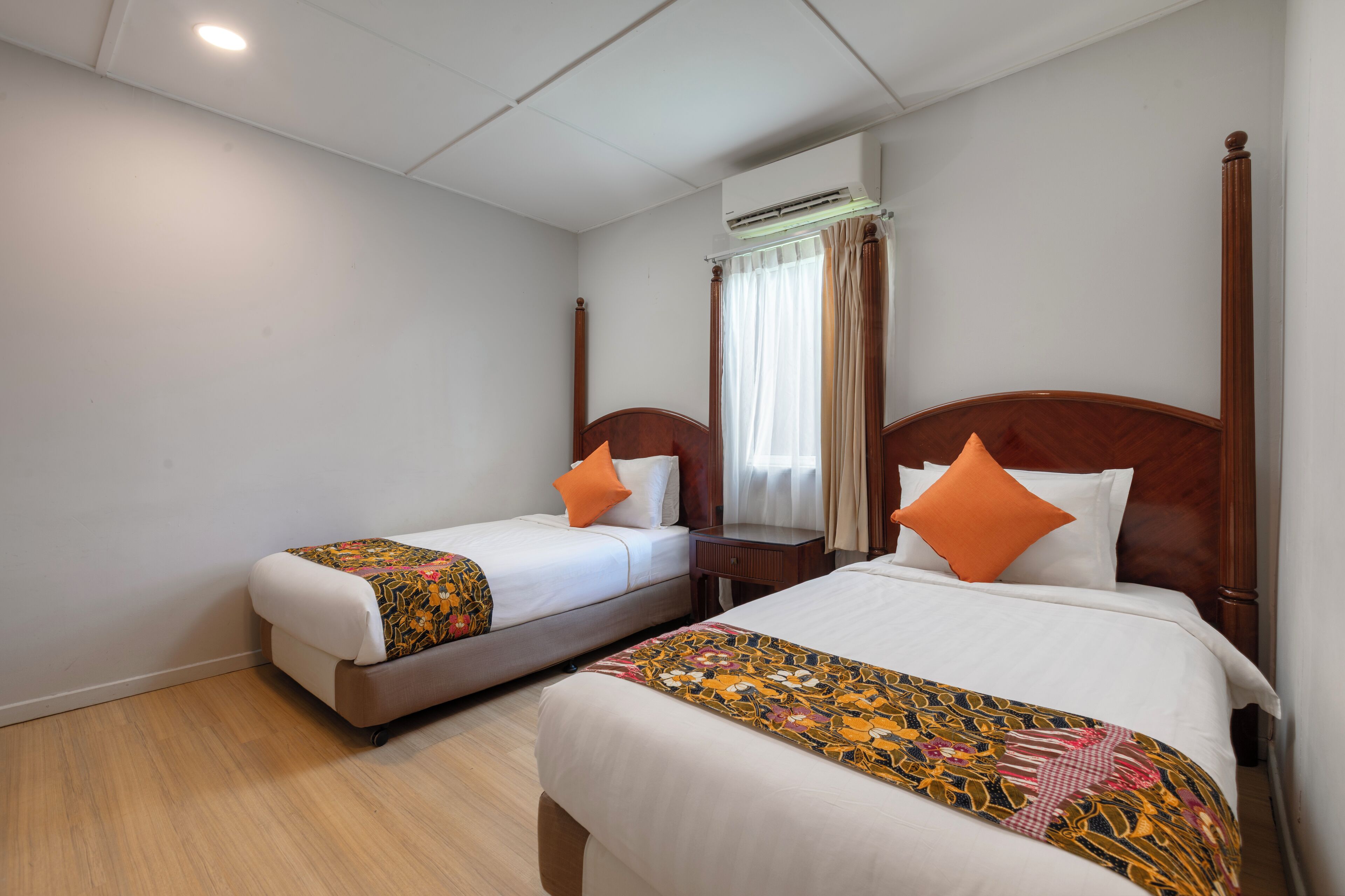 Hotelkamer met twee bedden, een houten hoofdbord, een nachtkastje, een airconditioning en een raam met gordijnen.