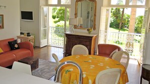 A living room with a sofa, dining table, chairs, a TV, and a balcony with a view of greenery. - 1st Floor Apartment With Beautiful Sea View in Magnificent Character Building (Cannes)