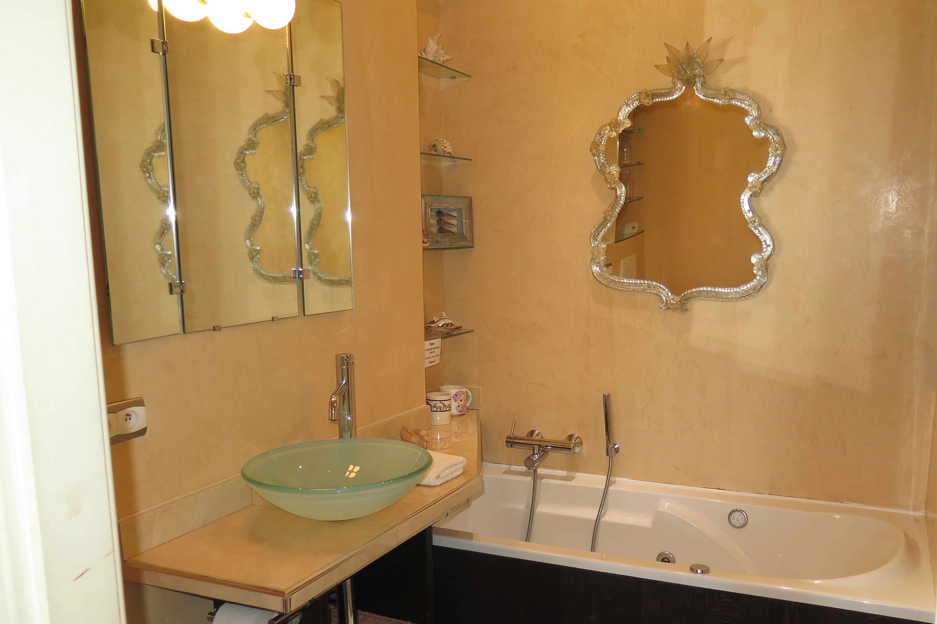 Une salle de bain dotée d’un lavabo en forme de bol en verre, d’une baignoire sur pieds et d’un miroir décoratif orné de motifs floraux.