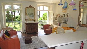 A spacious living room with a large window, a wooden cabinet, a sofa, a dining table, and a bookshelf. - 1st Floor Apartment With Beautiful Sea View in Magnificent Character Building (Cannes)