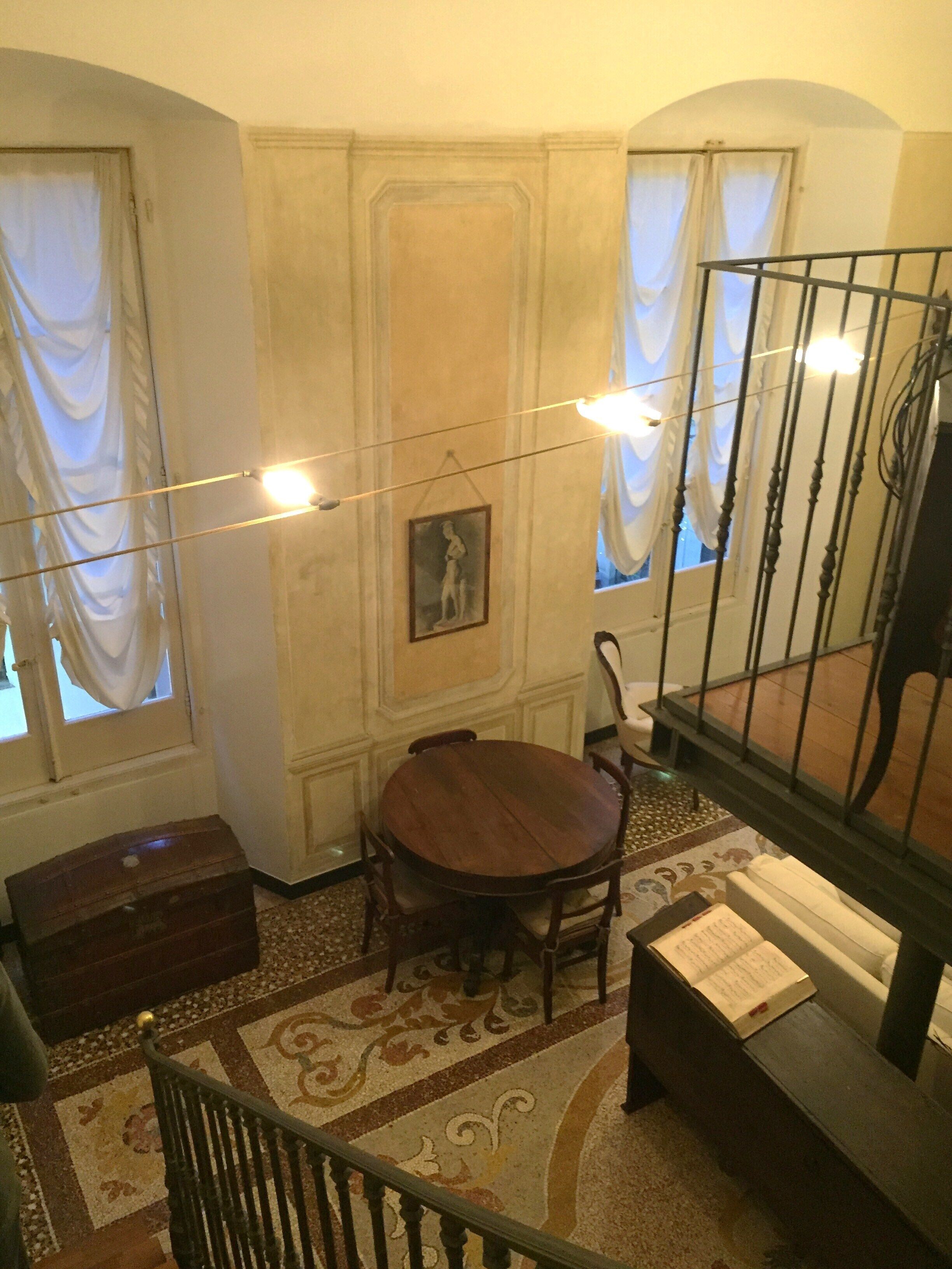 Una habitación con una mesa redonda de madera, una silla, un pequeño cofre de madera y una escalera con barandilla de metal.