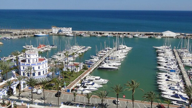 Un port de plaisance où sont amarrés de nombreux bateaux, un bâtiment à la façade bleue et blanche, et une vue dégagée sur la mer.
