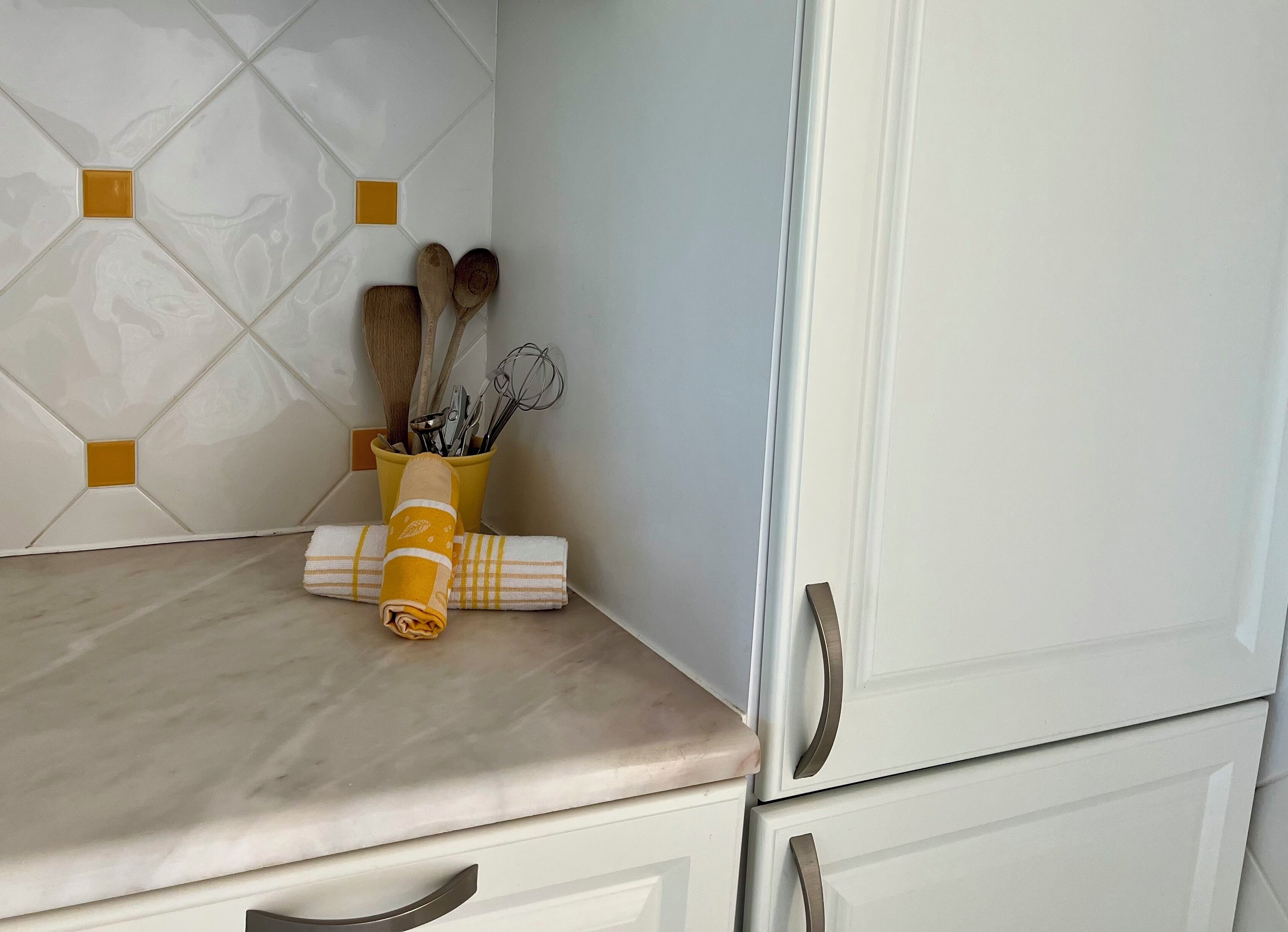 Un plan de travail de cuisine surmonté d’un plateau en marbre, avec une serviette jaune et un porte-ustensiles contenant des cuillères en bois et des fouets.