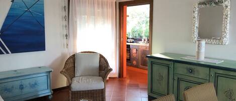 Una habitación acogedora con una silla de mimbre, una mesita con una taza, un armario verde y un cuadro en la pared.
