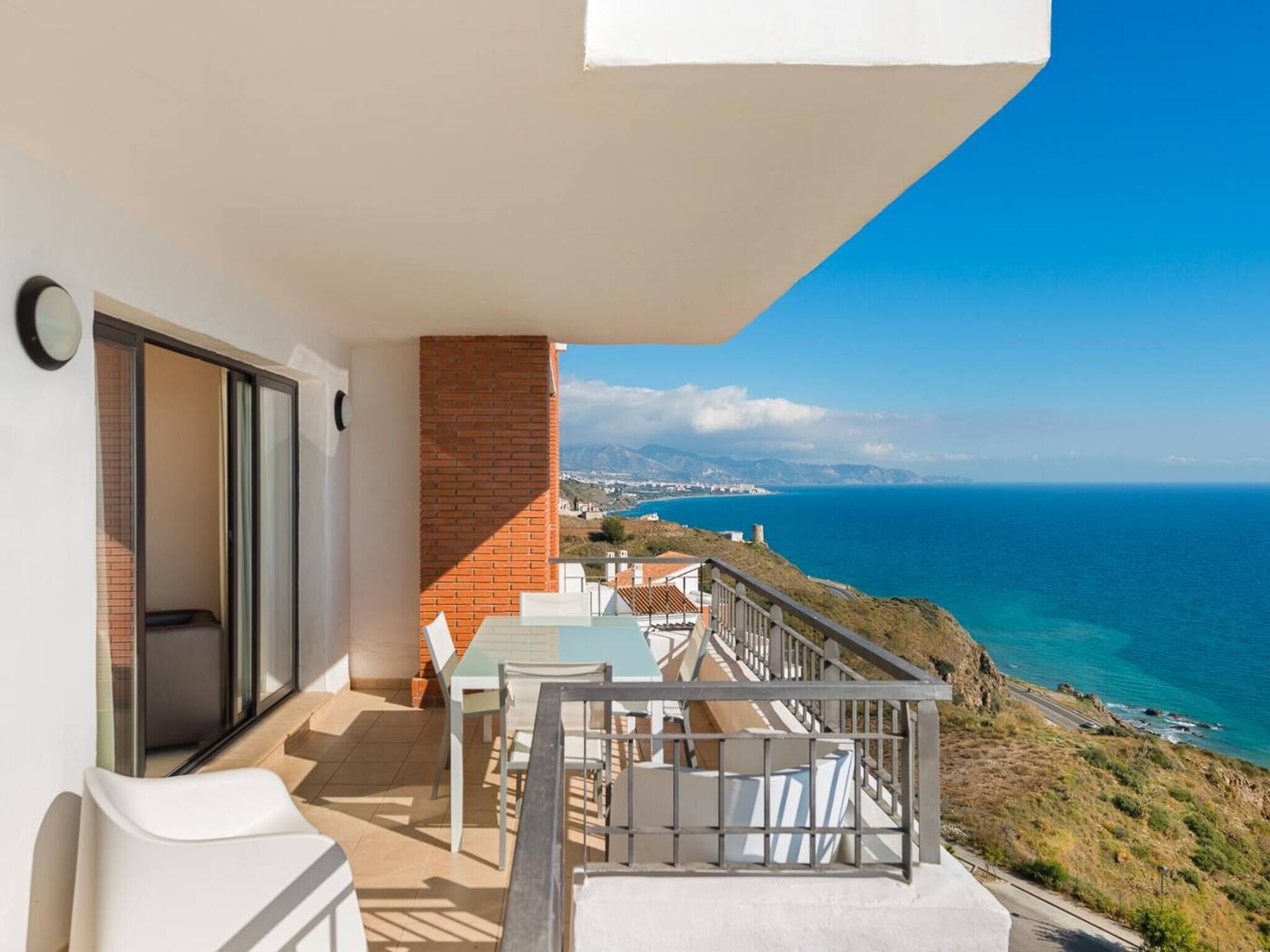 A balcony with a table and chairs, a brick wall, and a view of the sea and distant landscape.