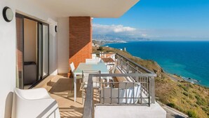 Un balcón con mesa y sillas, un muro de ladrillo y vistas al mar y el paisaje distante.